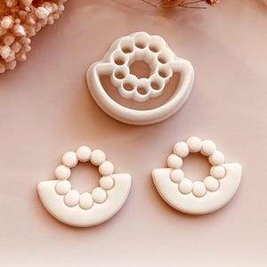 Puede incluir: Tres cortadores de arcilla polimérica blanca para manualidades. Los cortadores tienen forma de semicírculo con diseño de cuentas y forma circular con diseño de cuentas. Los cortadores están sobre una superficie rosa con flores secas.