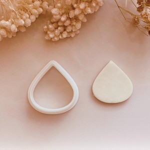 May include: Two teardrop-shaped cookie cutters. One is a white outline, and the other is a solid beige shape. The cutters are on a light pink surface, with dried flowers in the background. The image is from Babycutters, 2025.