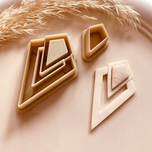 May include: Three geometric-shaped cookie cutters in beige plastic. The cutters are in the shape of a diamond with layered geometric shapes. The cutters are on a light pink surface. The brand name "Bezzycutters" is visible.