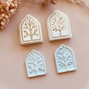 May include: Four white plastic cookie cutters in the shape of arched windows with tree designs. Two are white, and two are light blue. The cutters are on a pink plate with dried floral accents.