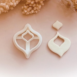 May include: Two white, ornate cookie cutters in an elegant teardrop shape, one with a cut-out design and the other a solid shape. The cutters are on a light pink surface, with dried flowers in the background. The image is from Babycutters, 2025.