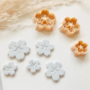 Puede incluir: Un conjunto de cortadores de galletas con forma de flor de color beige claro y varios recortes de fondant con forma de flor de color gris claro. Los cortadores son de plástico y tienen un diseño floral detallado. Los recortes son de varios tamaños.