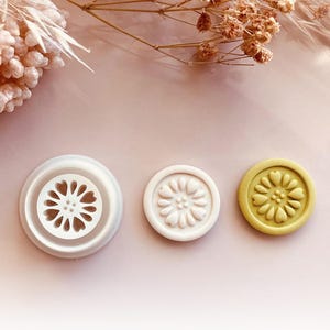 Peut inclure: Trois emporte-pièces ronds en forme de fleurs, blancs et vert-jaune. Le plus grand a un motif découpé, les autres ont des motifs floraux en relief. Le fond est rose clair avec des fleurs séchées.