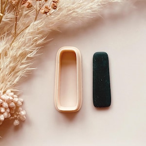 May include: A light pink rectangular cookie cutter next to a dark green rectangular piece of clay. The cookie cutter has a rounded top and bottom. The background is a light pink surface with dried flowers.