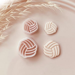 Op de afbeelding: Vier volleybalvormige koekjessnijders en koekjes. Twee zijn lichtroze plastic snijders en twee zijn afgewerkte koekjes. De koekjes en snijders liggen op een lichtroze ondergrond met gedroogd gras.