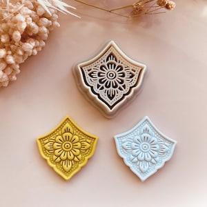 May include: Three decorative cookie cutters in a floral design. One is gray, one is yellow, and one is light blue. The cutters are diamond-shaped with a detailed flower pattern in the center. The image includes the text "Babycutters, 2025".