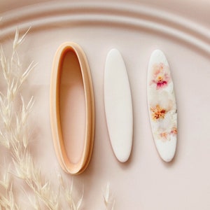 Könnte beinhalten: Ein Set aus drei ovalen Ausstechern. Einer ist hellpfirsichfarben, die anderen beiden sind weiß, einer mit floralem Design. Das Bild befindet sich auf einer hellrosa Oberfläche mit getrocknetem Gras.