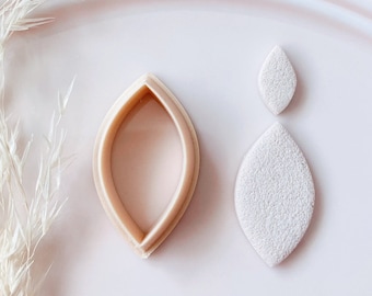 May include: A light beige polymer clay earring cutter and two clay earring pieces. The cutter is in the shape of an elongated oval. The clay pieces are white with a textured surface. The background is a soft pink.