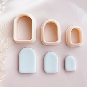 May include: A collection of six arch-shaped polymer clay cutters and clay pieces in different sizes. The cutters are a light peach colour, and the clay pieces are a pale blue. They are arranged on a pink surface.