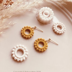 Puede incluir: Pendientes y piezas decorativas de arcilla polimérica hechos a mano. Los pendientes tienen forma de girasol, con un centro amarillo y un anillo exterior blanco. Las piezas están dispuestas sobre una superficie rosa claro con flores secas.