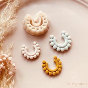 Puede incluir: Vista cenital de cuatro cortadores de galletas en forma de U en blanco roto, azul claro y amarillo, decorados con pequeños detalles redondos. La imagen está sobre un plato rosa, con flores secas en el fondo.