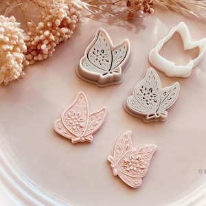 May include: A collection of butterfly-shaped cookie cutters in various shades of pink and gray, with intricate floral and leaf patterns. One white cutter is also present. The cutters are arranged on a light pink plate, with dried floral accents.