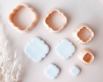 Puede incluir: Un conjunto de cortadores de galletas en forma de cuadrifolio rosa claro, de varios tamaños, junto con varios recortes de fondant cuadrifolio azul claro. Los artículos están dispuestos sobre un plato rosa claro, lo que sugiere un tema de repostería o manualidades.