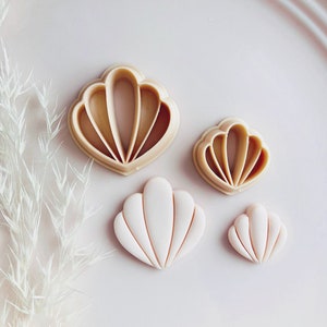 Può includere: Quattro formine per biscotti a forma di conchiglia di diverse dimensioni. Due sono in plastica beige e due sono bianche. Le formine sono disposte su una superficie rosa chiaro.