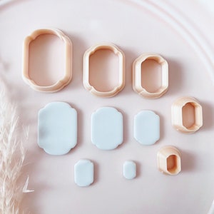 May include: A collection of eight beige-colored, decorative, geometric-shaped cookie cutters and six light blue, geometric-shaped cutouts. The cutters are arranged on a light pink surface, with a feathery plant in the background.