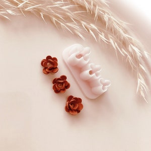 Puede incluir: Un conjunto de tres pequeñas decoraciones en forma de flor de color óxido y un cortador de flores de plástico blanco. Las flores tienen pétalos detallados y un pequeño centro brillante. El cortador tiene múltiples moldes con forma de flor.