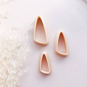 May include: Three light pink, teardrop-shaped cookie cutters of varying sizes. The cutters are arranged on a light pink surface, with a white feathery plant in the background. The cutters have a smooth, rounded edge.