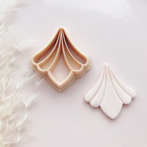 May include: Two beige, fan-shaped cookie cutters on a pink surface. One cutter is a plastic mold, and the other is a baked cookie. The cookie cutter has a scalloped edge and a detailed design.