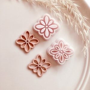 Może przedstawiać: Cztery różowe wykrawacze do ciastek w kształcie kwiatów na jasnoróżowym talerzu. Dwa wykrawacze są pełne, a dwa mają wycięty wzór. Obraz zawiera suszoną trawę ozdobną i tekst "Copyright © 2023 Babycutters."