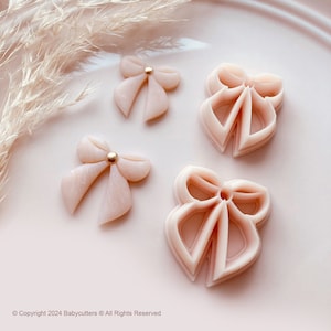 Puede incluir: Cuatro cortadores de galletas en forma de lazo rosa claro con un centro de cuentas doradas y dos lazos terminados. Los lazos están dispuestos en un plato rosa con hierba ornamental seca. Copyright 2024 Babycutters All Rights Reserved.