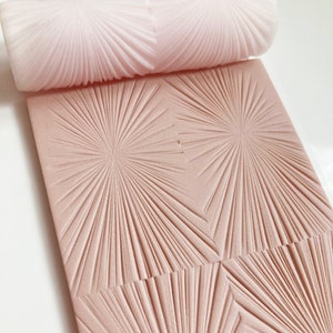 May include: A light pink textured rolling pin and a matching rectangular surface with a radiating starburst pattern. The rolling pin is cylindrical, and the surface is a rectangle with a repeating design. The image is a close-up shot.