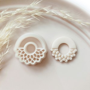 Pode incluir: Dois cortadores de biscoitos em forma de flor, em branco sujo. Um é um semicírculo com um design detalhado de pétalas, e o outro é um círculo inteiro menor com um padrão de pétalas semelhante. Os cortadores estão em uma superfície rosa claro.