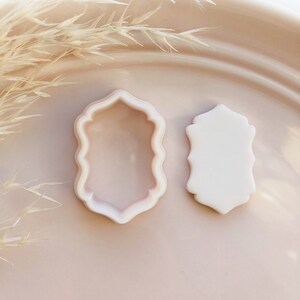 May include: Two light pink cookie cutters in a decorative shield shape, one a cutter and the other a solid shape. The cutters are on a light pink plate with dried ornamental grass in the background. The copyright notice is at the bottom.