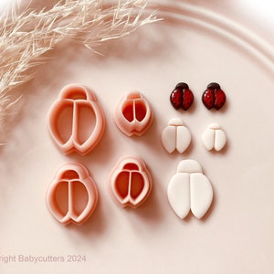 Puede incluir: Un conjunto de cortadores de galletas y decoraciones de fondant con temática de mariquita. Los cortadores son de color rosa claro, y las piezas de fondant son blancas, rojas y negras. La imagen incluye el texto "Copyright Babycutters 2024".