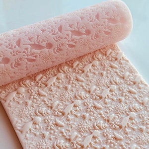 Op de afbeelding: Een lichtroze deegroller en een bijpassende rechthoekige mat, beide met een herhalend patroon van vogels, bloemen en bladeren. De deegroller is cilindrisch en rust op de mat. Het patroon is in reliëf.