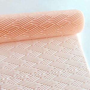 Può includere: Un set di mattarello e tappetino color pesca chiaro con un motivo intrecciato. Il mattarello è cilindrico e arrotolato sul tappetino rettangolare. Il tappetino e il mattarello sono realizzati in materiale flessibile. Il testo "BABYCUTTERS 2024" è in basso a destra.