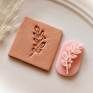 May include: A terracotta-coloured square stamp and a pink oval stamp, both featuring a botanical design of leaves and berries. The square stamp has the design pressed into the surface, the oval stamp has the design raised.