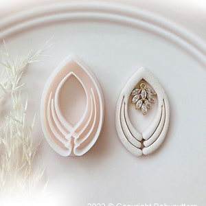 Peut inclure: Deux emporte-pièces en pâte polymère en forme de goutte d'eau, l'un avec un charm doré orné de cristaux transparents. Les emporte-pièces sont rose clair et blancs, avec un design en couches. L'image est sur fond blanc.