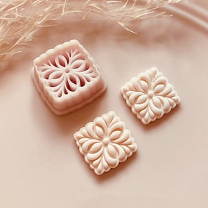 Puede incluir: Tres cortadores de galletas cuadrados de color rosa claro con diseños florales. Uno es un molde y dos son galletas terminadas. Los cortadores están sobre un plato rosa con hierba seca en el fondo. Copyright Babycutters 2024.