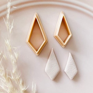 May include: A collection of diamond-shaped polymer clay cutters and finished clay earrings. The cutters are light brown, and the earrings are a textured off-white. The items are arranged on a light pink plate with dried decorative grass.