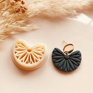 May include: A beige polymer clay earring and matching cutter. The earring is a dark teal color with a gold-colored stud and accent. The cutter is a light beige color with a floral design. The image also includes dried flowers.