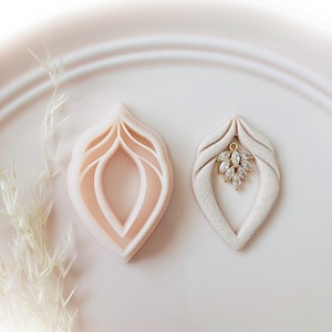 May include: Two leaf-shaped polymer clay cutters, one hollow and the other with a gold-colored charm with clear crystals. The cutters are light pink and off-white, set on a white plate. Copyright © 2022 Babycutters is printed at the bottom.