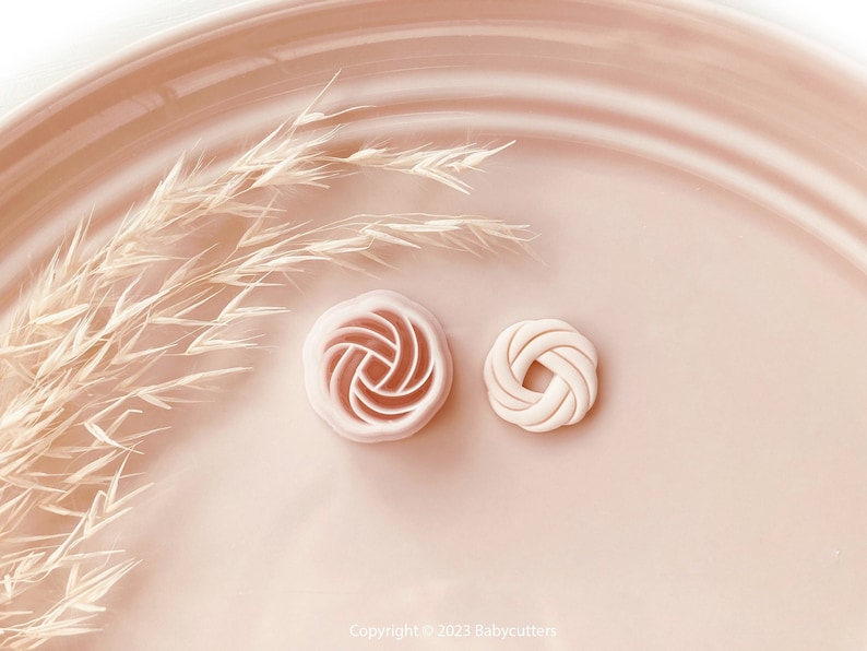 Puede incluir: Dos cortadores de galletas de color rosa claro, uno con forma de rosa y otro de pretzel, sobre un plato rosa a juego. Hierba beige seca acent&uacute;a la escena. Se ve el aviso de copyright "Copyright &copy; 2023 Babycutters".