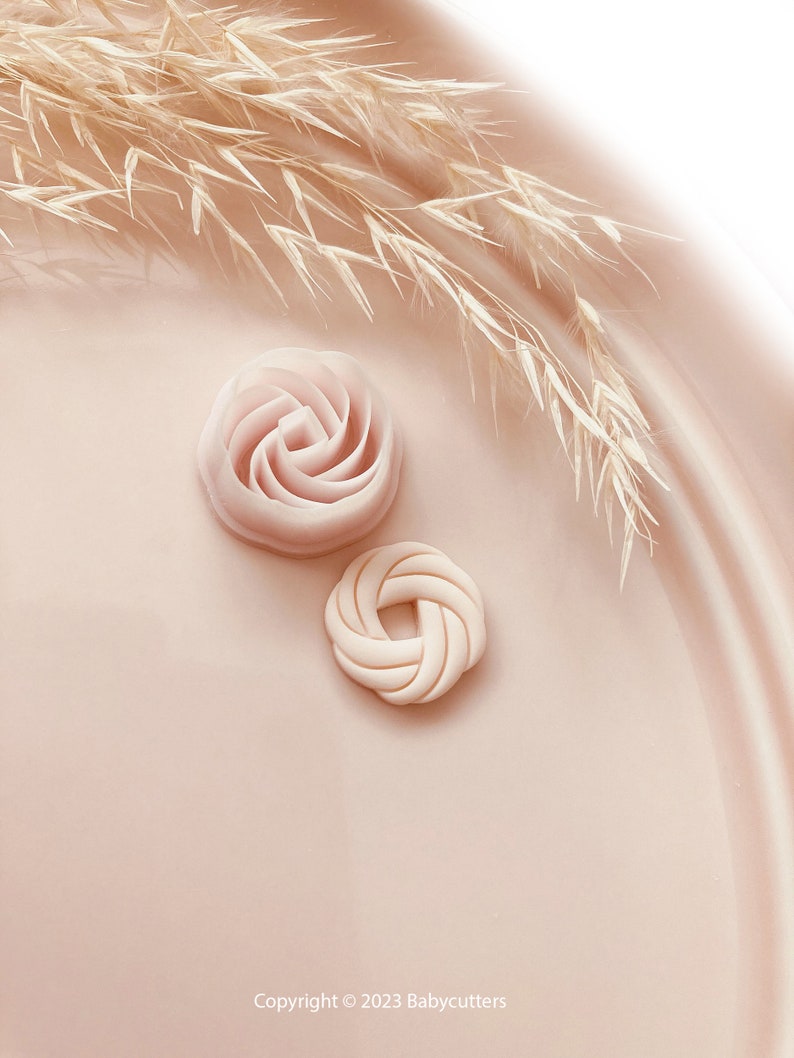 Puede incluir: Dos objetos rosas en forma de flor y hierba ornamental seca sobre una superficie rosa claro. Un objeto tiene forma de rosa y el otro de nudo. Los objetos son probablemente para manualidades o reposter&iacute;a.
