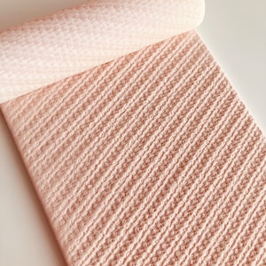 May include: A rolled-up, textured, light pink material with a diagonal pattern. The material is partially unrolled, revealing a detailed surface. The image is well-lit, highlighting the texture and colour of the material. The bottom left corner of the image has the text "@babycutters".