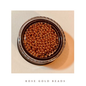 May include: A clear plastic container filled with numerous small, rose gold beads. The container has a black rim and a clear lid. The words "ROSE GOLD BEADS" are printed in a serif font below the container.