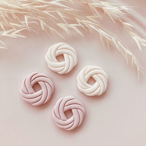 Puede incluir: Cuatro objetos decorativos circulares, blanquecinos y rosas, con un dise&ntilde;o tejido, est&aacute;n dispuestos sobre una superficie rosa. Los objetos son de diferentes tonos de rosa y blanco. Plantas beige secas y plumosas en el fondo.