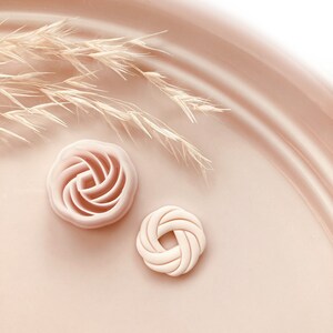 Puede incluir: Dos cortadores de galletas decorativos de color rosa claro. Uno tiene forma de rosa y el otro de nudo. Los cortadores est&aacute;n sobre una superficie rosa claro con unas ramitas de hierba seca en el fondo. Copyright 2023 Babycutters est&aacute; impreso en la parte inferior.
