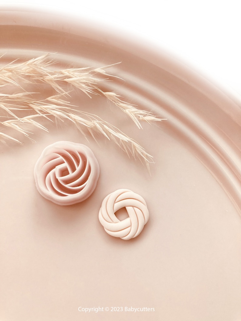 Puede incluir: Dos cortadores de galletas decorativos de color rosa claro. Uno tiene forma de rosa y el otro de nudo. Los cortadores est&aacute;n sobre una superficie rosa claro con unas ramitas de hierba seca en el fondo. Copyright 2023 Babycutters est&aacute; impreso en la parte inferior.