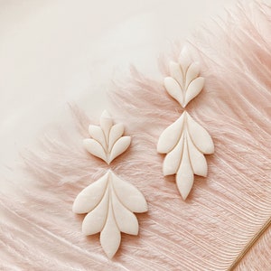 May include: Four off-white, leaf-shaped earrings are arranged on a pink surface with feather-like textures. The earrings have a detailed, carved design with multiple lobes. The earrings are in various sizes, with two pairs of matching shapes.