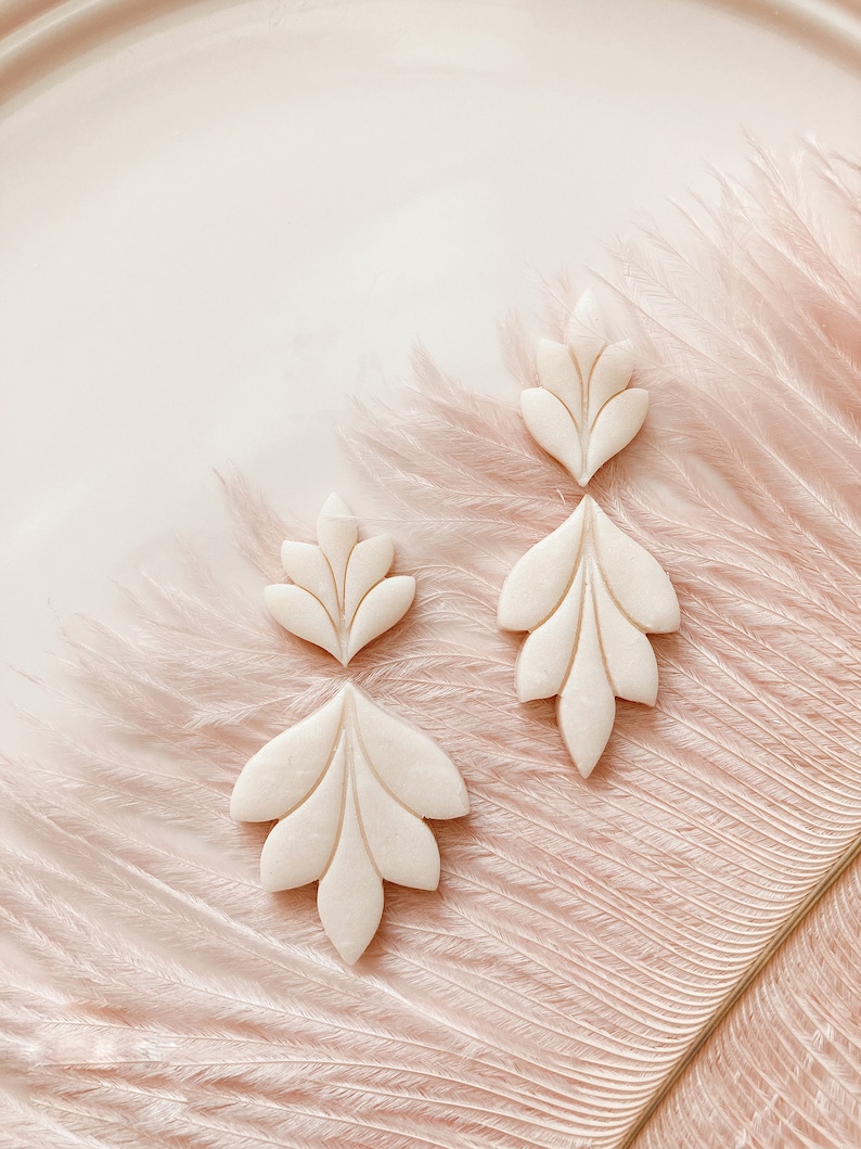 May include: Four off-white, leaf-shaped earrings are arranged on a pink surface with feather-like textures. The earrings have a detailed, carved design with multiple lobes. The earrings are in various sizes, with two pairs of matching shapes.