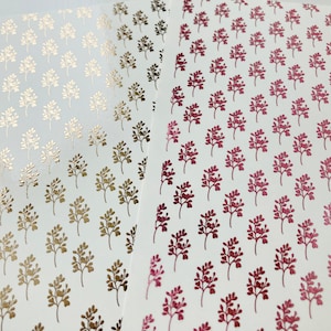 May include: Sheets of decorative stickers featuring a repeating pattern of small, stylized branches. The stickers are in gold and shades of pink and burgundy, arranged on a white background. The stickers are likely for crafts or scrapbooking.