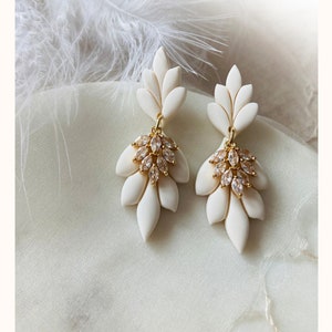 May include: Ivory-coloured, leaf-shaped earrings with gold-coloured detailing and crystal clusters. The earrings are displayed on a soft, white fabric. The designer's name, "Crown In The Jewels", is at the bottom of the image.