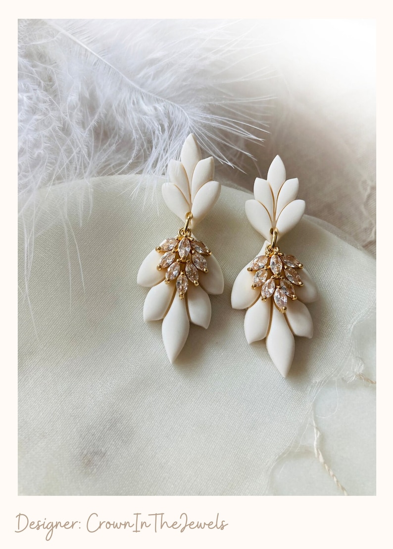 May include: Ivory-coloured, leaf-shaped earrings with gold-coloured detailing and crystal clusters. The earrings are displayed on a soft, white fabric. The designer's name, "Crown In The Jewels", is at the bottom of the image.