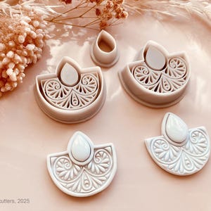 May include: A collection of light gray polymer clay cutters in a floral design. The cutters are in the shape of a half-circle with a teardrop shape on top. The cutters are for making earrings or other jewelry.
