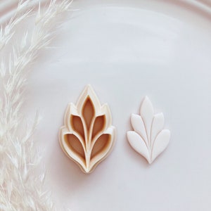 May include: Two leaf-shaped cookie cutters. One is beige and the other is white. The beige cutter has a detailed design, while the white one is a simpler version. The background is a white plate with white decorative elements.